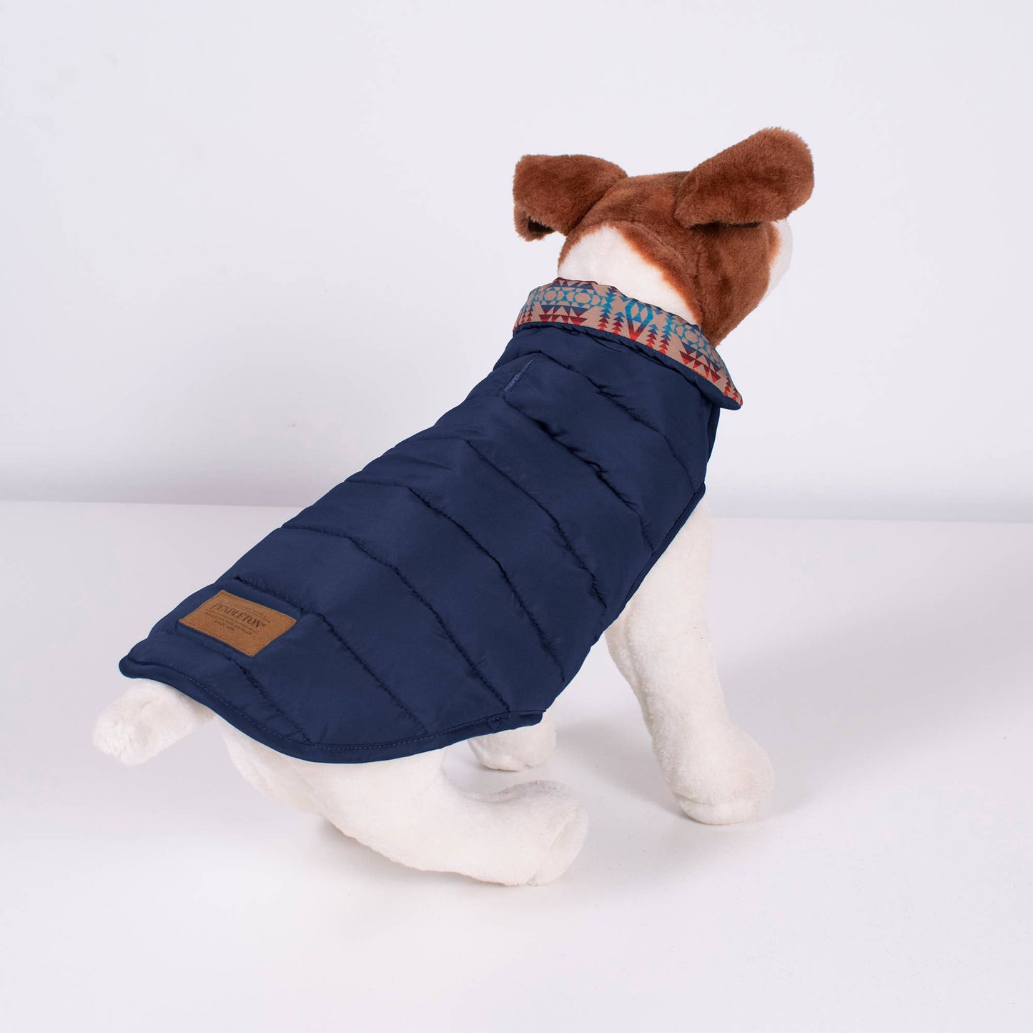 A durable and insulated dog coat in a sleek puffer style, featuring a rich Pilot Rock pattern with earthy tones of gray, navy, and hints of red. The coat is designed with adjustable straps for a secure and comfortable fit, providing warmth and protection for your pet in cold weather. The quilted design offers both function and a stylish, rugged look, perfect for outdoor adventures.